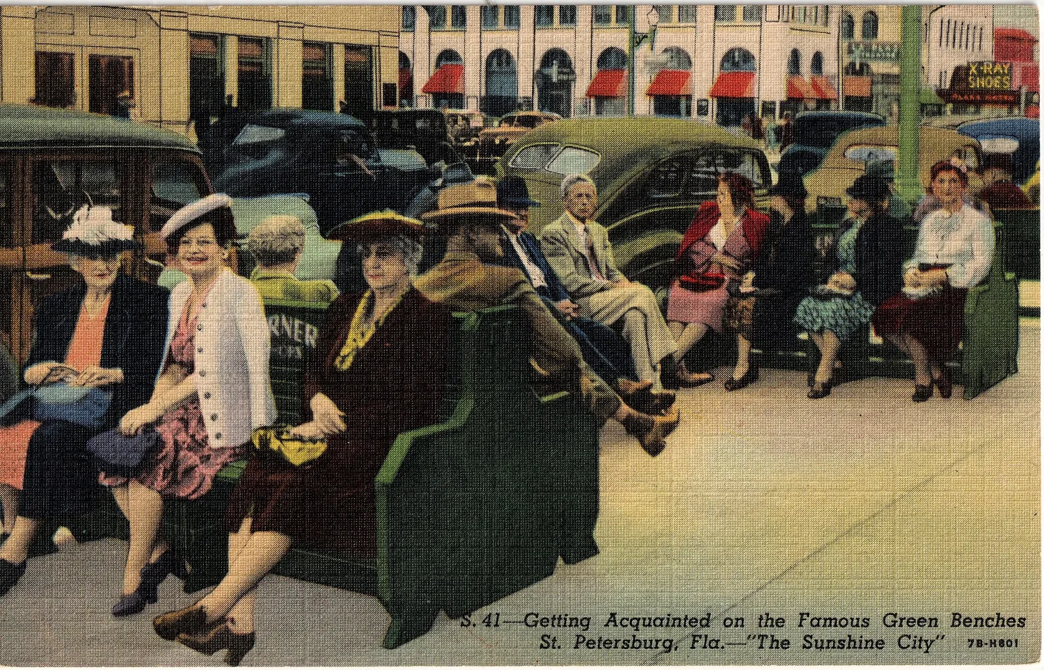 People of different ages sitting close together and chatting on the green benches, 1940s, storefronts and parked cars behind them.