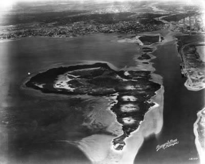 A 1925 aerial photograph of two small, flat, marshy islands surrounded by open bay water, before any construction.