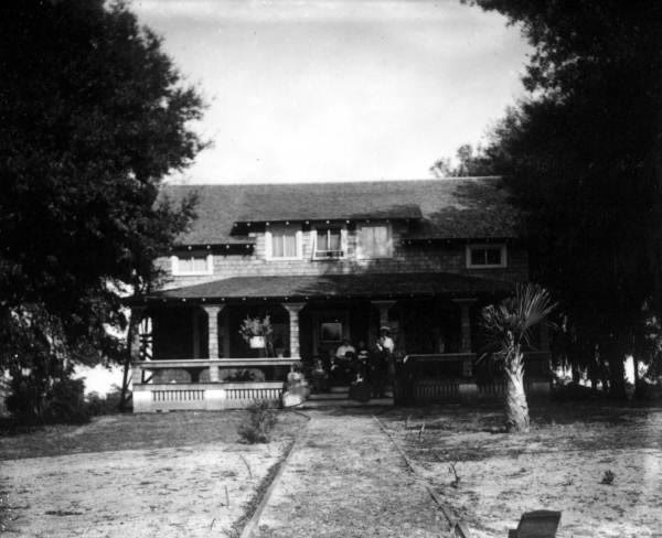 A house with a deep front porch under shade trees; several family members sit together on the porch, with a sandy path leading up to it.