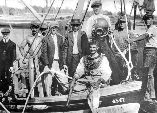 A diver in a canvas suit sits in a small boat holding an air hose, surrounded by about eight men in caps and jackets; a bronze diving helmet is held up beside him.