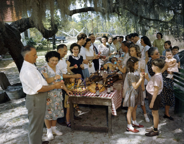 A large family gathered under mossy oak trees around a table crowded with food and bottles, several people holding up dyed red eggs to knock together, in a color photograph.