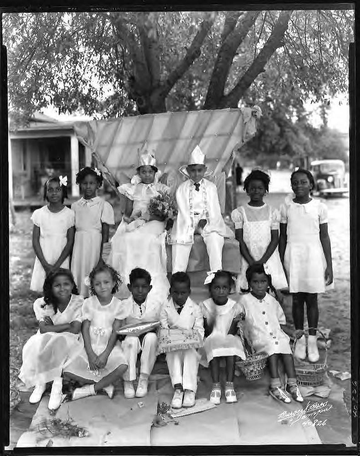 The May Day King and Queen of Dobyville School and their court, a dozen Black schoolchildren seated under a tree, 1937.